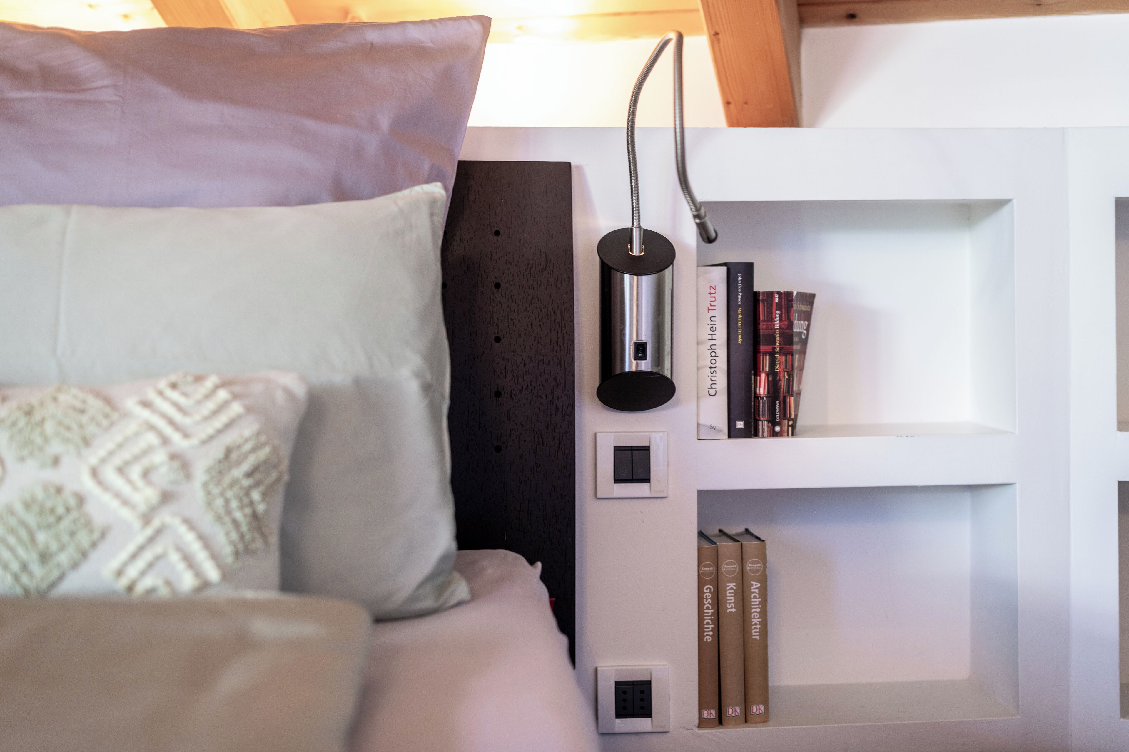 A bedroom with a bed, pillows, a bedside table, a wall-mounted shelf with books, and a lamp.
