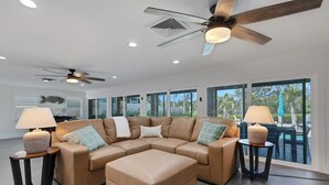 A spacious living room with a large L-shaped sectional sofa, two side tables with lamps, ceiling fans, and large windows offering a view of palm trees.