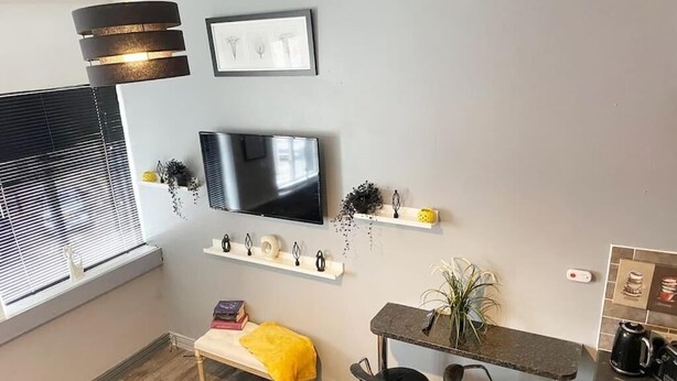 A modern living room with a flat-screen TV mounted on the wall, a bench with a yellow throw, and a small bar area with stools. - The Anchor - Open Plan Living in South Shields (South Shields)