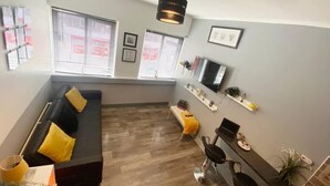 A modern living room with a black sofa, yellow cushions, a coffee table, a TV mounted on the wall, and a small desk with a chair. - The Anchor - Open Plan Living in South Shields (South Shields)
