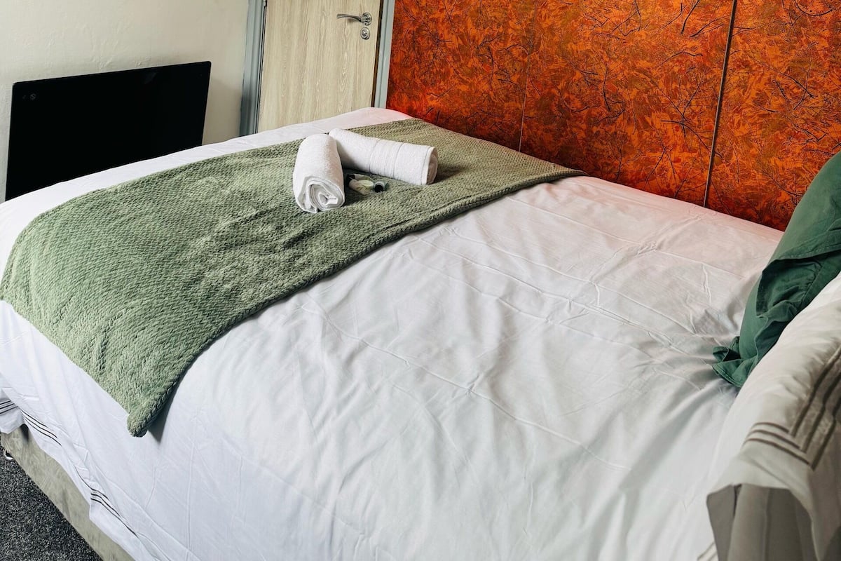 A hotel room with a bed, a green blanket, a white towel, a wooden door, and a mirror on the wall.