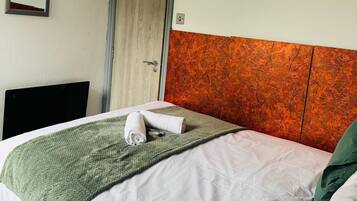 Quarto de hotel com uma cama, um cobertor verde, uma toalha branca, uma porta de madeira e um espelho na parede.