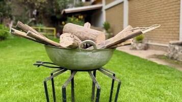 Ein Metallgrill mit Holzscheiten auf einer grünen Rasenfläche, umgeben von Bäumen und einem Gebäude im Hintergrund.