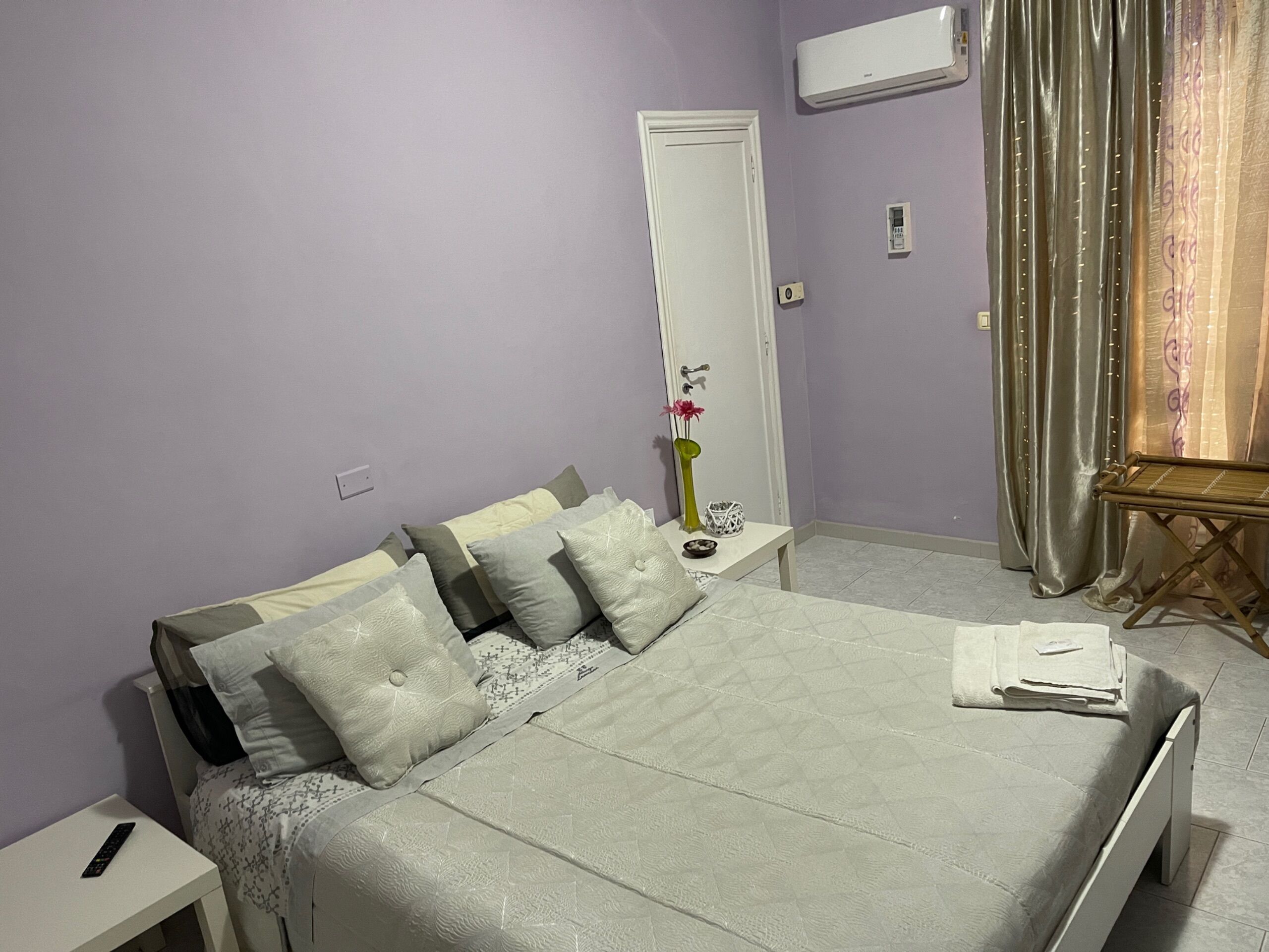 A neatly made bed with pillows, a bedside table with a remote control, a small table with a vase and a decorative item, and a window with curtains.