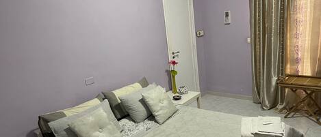 A neatly made bed with pillows, a bedside table with a remote control, a small table with a vase and a decorative item, and a window with curtains.