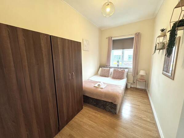 Une chambre avec une grande armoire, un lit recouvert d’une couverture rose, une fenêtre avec des rideaux et une petite table de chevet avec une lampe.