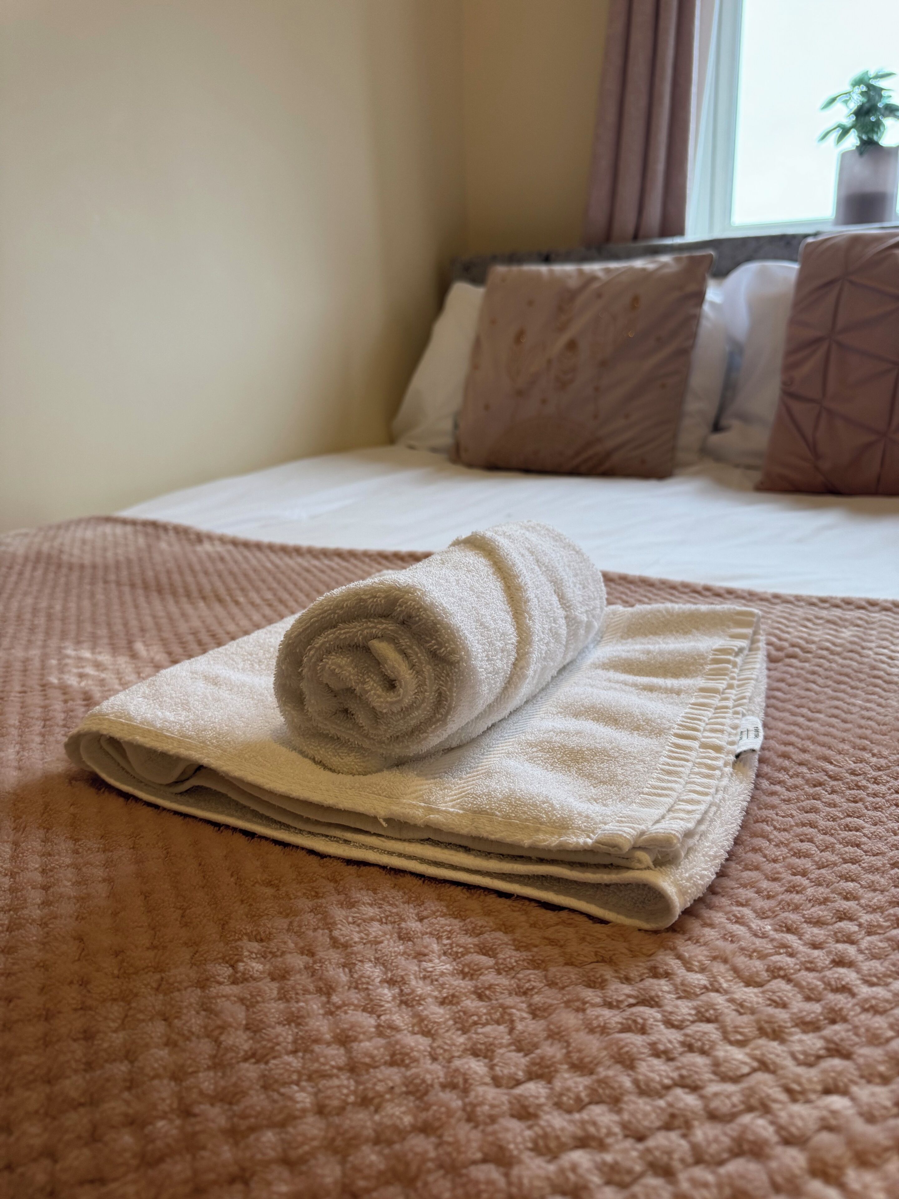 A neatly made bed with a rolled-up towel and a folded blanket on a textured pink bedspread.