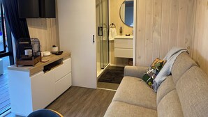 Ett modernt vardagsrum med en grå soffa, ett TV-bord i trä, en liten fågelbur och en skjutdörr som leder till ett badrum med spegel och handfat.