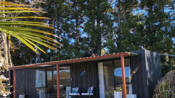 En modern stuga med en veranda, stora fönster och en liten sittplats utomhus.