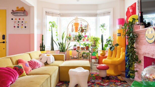 Un salón vibrante con un sofá amarillo, cojines coloridos, un taburete con forma de diente y una variedad de plantas y juguetes.
