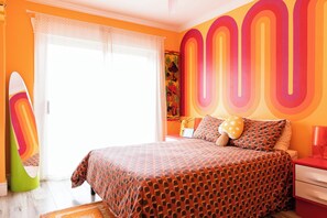 Une chambre avec un lit, une table de chevet, une lampe et une planche de surf appuyée contre le mur.