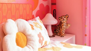 Un dormitorio con pared rosa, cama con cubrecama floral, un peluche de leopardo, una lámpara blanca y una planta colgando del techo.