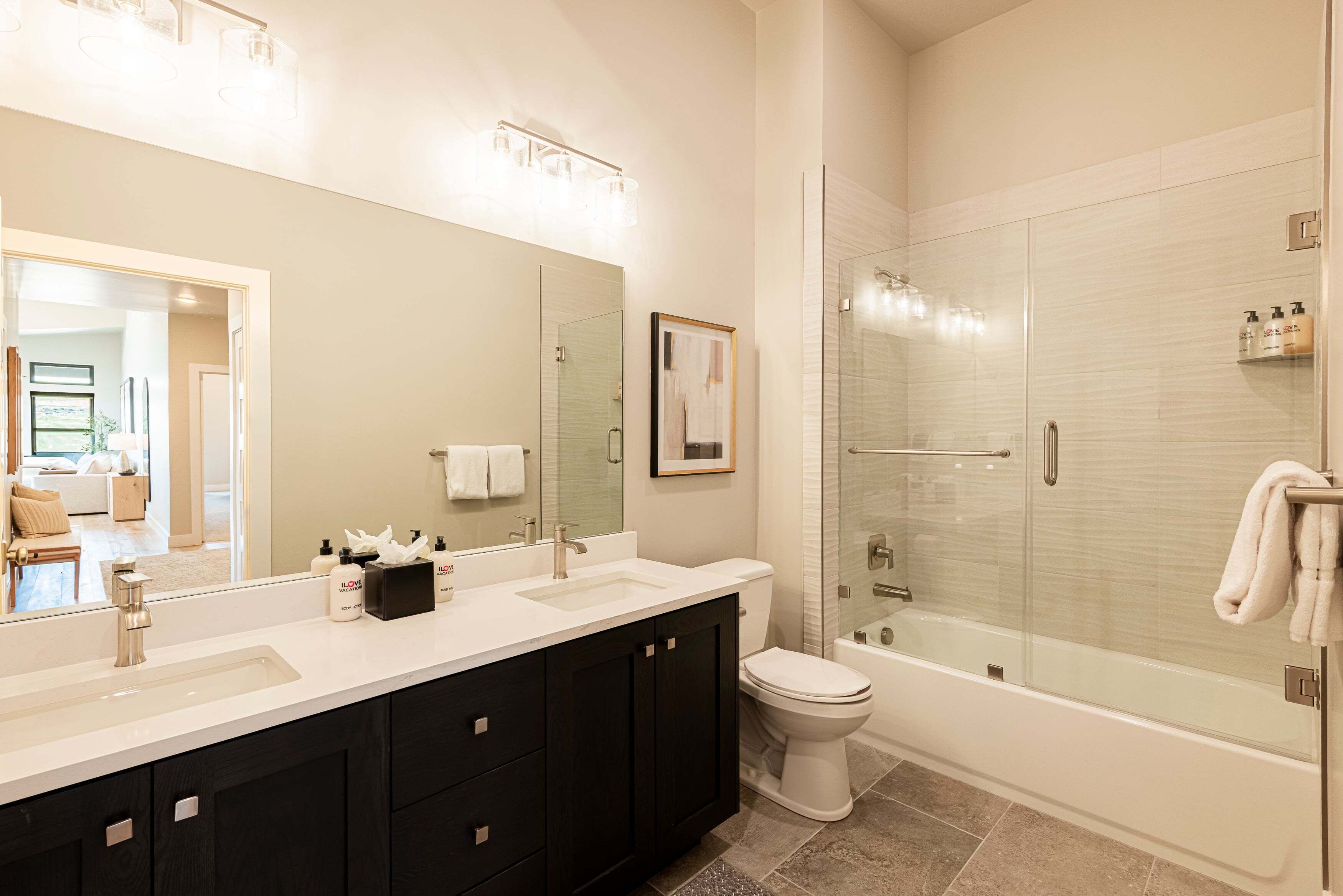 A modern bathroom with a large mirror, dual sinks, dark cabinets, a toilet, and a glass-enclosed shower area.
