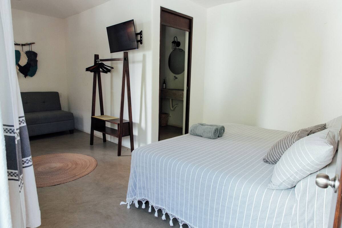 Un dormitorio con cama, un sofá gris, un televisor sobre un mueble y la puerta del baño.