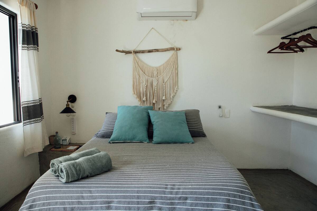 Una cama bien tendida con ropa de cama a rayas, dos almohadas y una toalla enrollada. Un colgador de macramé está sobre la cama, y un perchero a la derecha.
