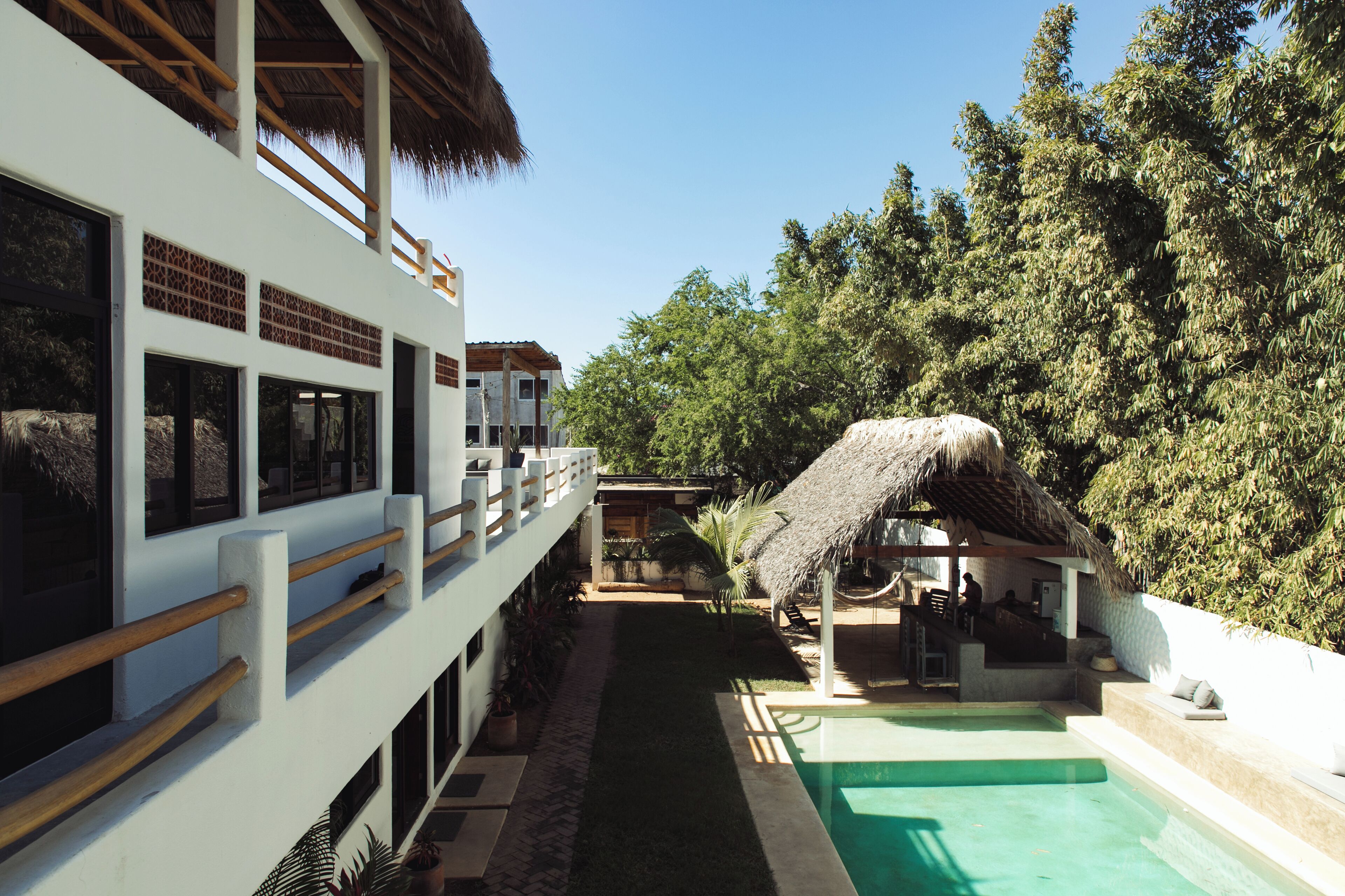 Un área de piscina con una estructura de techo de paja, un edificio blanco con ventanas amplias y abundante follaje.