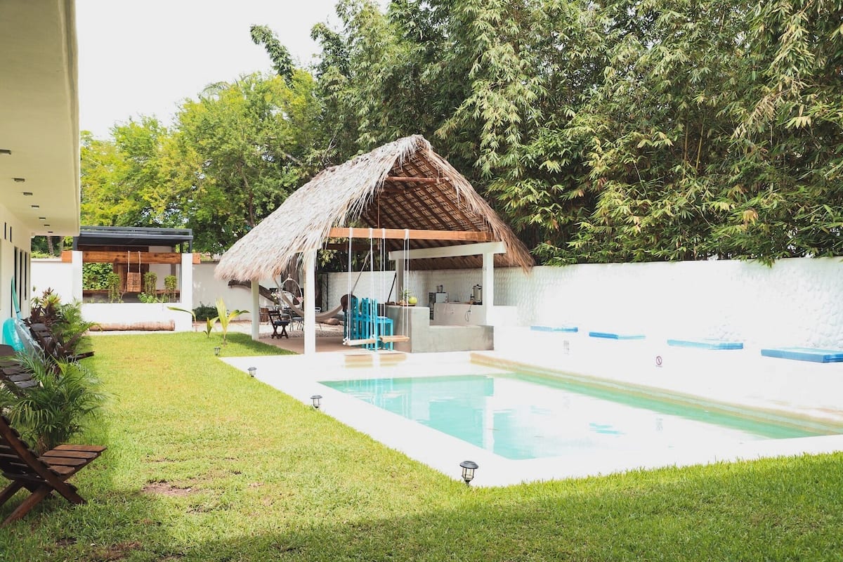 Un área de piscina moderna al aire libre con un pabellón de techo de paja, un césped y una pared blanca.