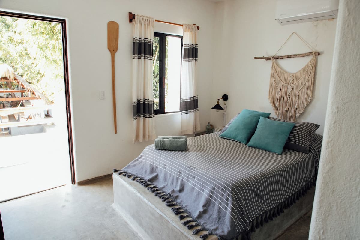 Un dormitorio con una cama, una ventana con cortinas, un tapiz de macramé en la pared y una paleta de madera colgada.