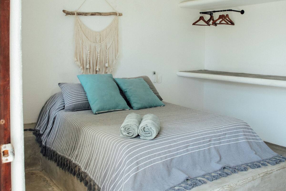 Un dormitorio con una cama, ventilador de techo, unidad de aire acondicionado, un colgador de macramé y una estantería con perchas.
