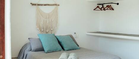 A bedroom with a bed, a ceiling fan, an air conditioning unit, a macrame wall hanging, and a shelf with hangers.
