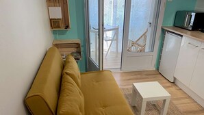 A cozy room with a mustard-colored sofa, a small white table, and a kitchen area with white cabinets and a microwave. - Le Cocon de la corniche St Hilaire (SAINT HILAIRE DE RIEZ)