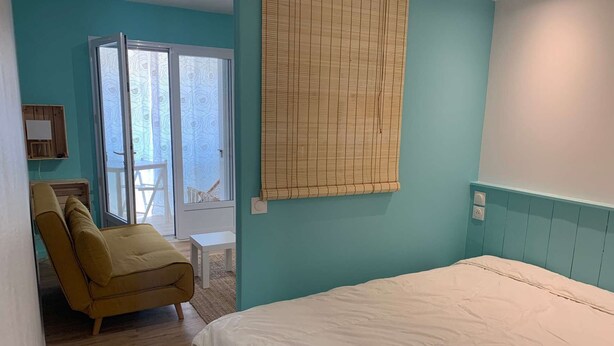 A bedroom with a bed, a chair, a small table, and a window with blinds. - Le Cocon de la corniche St Hilaire (SAINT HILAIRE DE RIEZ)