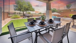 An outdoor patio with a table set for dining, surrounded by chairs, and a view of a backyard with a pool and barbecue area. - Remodeled 4BR Queen Creek Home | Pool, Game Room, Fire Pit (Queen Creek)