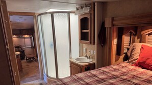 A bedroom with a plaid bedspread, a bathroom with a sink and mirror, and a shower area. - Cozy 1-bedroom Rv with pool,WiFi, AC in pleasant Elephant Butte (Elephant Butte)