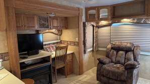 A compact RV kitchen with wooden cabinets, a flat-screen TV, a dining area with a chair, and a recliner. - Cozy 1-bedroom Rv with pool,WiFi, AC in pleasant Elephant Butte (Elephant Butte)