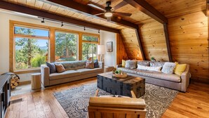 A cozy loft-style room with a sloped ceiling, large windows offering a view of trees, a fireplace, and two sofas with cushions.
