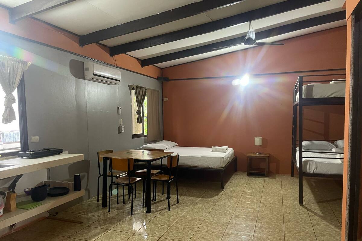 A compact room with a bunk bed, a desk with a chair, a small table, and a bed with a white mattress.
