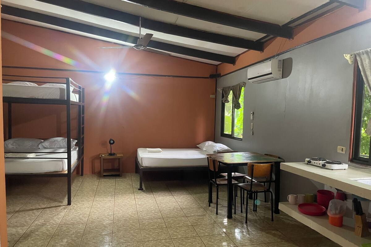 A dormitory room with bunk beds, a single bed, a desk with chairs, a small table, a ceiling fan, an air conditioning unit, and a window with curtains.