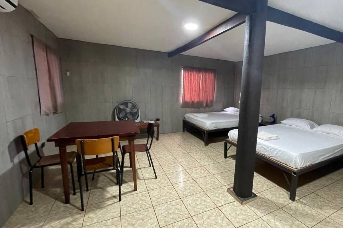 A room with a wooden table and chairs, two single beds, a standing fan, and a ceiling fan.