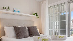 A hotel room with a bed, two pillows, a nightstand with a lamp, a window with a view, and a shelf with plants and a painting. - Santana | Sea views - garden - terrace (La Cala de Mijas)