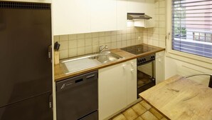 A compact kitchen with a refrigerator, dishwasher, oven, and a window with blinds.