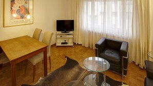 A living room with a wooden dining table, a leather sofa, a glass coffee table, a flat-screen TV on a stand, and a large window with sheer curtains.