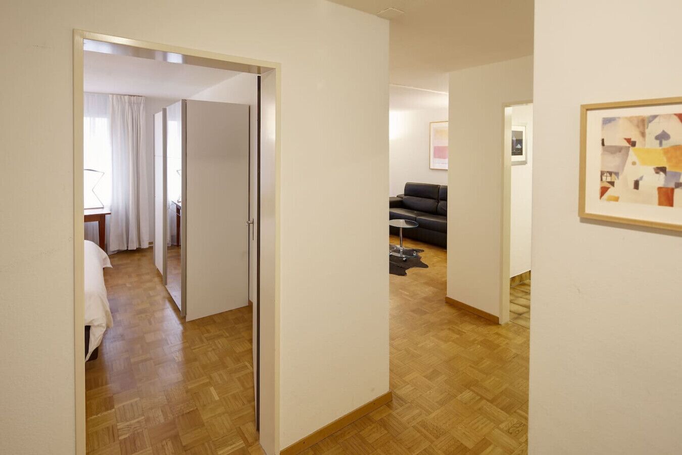 A hallway with a wooden floor leading to a room with a bed and a window with curtains. There is a door on the left and a framed picture on the right wall.