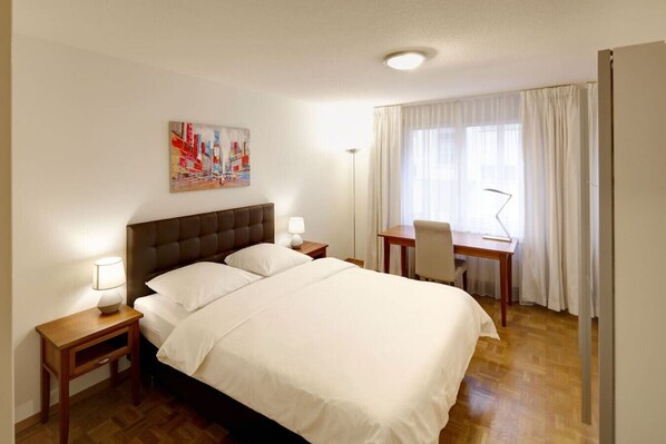 A hotel room with a large bed, a desk, and a chair. There is a painting on the wall and a window with curtains.