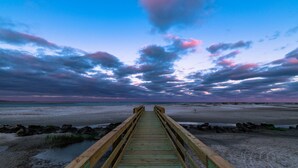 Eine Holzstege führt zu einem Strand, der von einem klaren Himmel mit Wolken gesäumt ist.