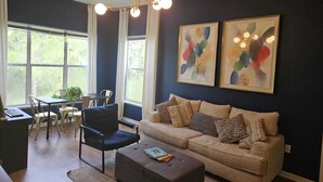 Ein modernes Wohnzimmer mit dunkelblauer Akzentwand, einem beigen Sofa, einem schwarzen Sessel, einem Essbereich mit Tisch und Stühlen sowie zwei abstrakten Gemälden.