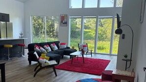A spacious living room with a large window, a black sofa, a red rug, and a chess table.