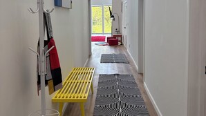 A hallway with a coat rack, a yellow bench, patterned rugs, and a view of a red chair and a window with natural light.