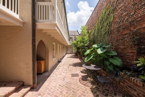 A narrow courtyard with brick flooring, a white building with balconies, and potted plants along the brick wall.