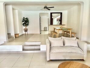 A modern living room with a sofa, a dining table with benches, a ceiling fan, and a staircase leading to another level.