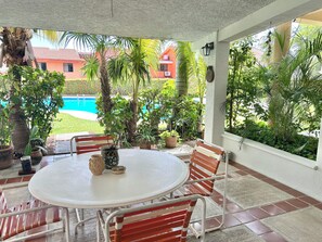 Un porche techado con una mesa redonda y sillas, plantas en macetas y vistas a una piscina y edificios.
