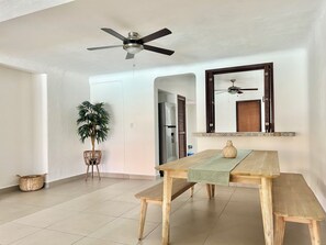 Un coin repas avec une table en bois, des chaises, un ventilateur de plafond, une plante en pot et un miroir au mur.