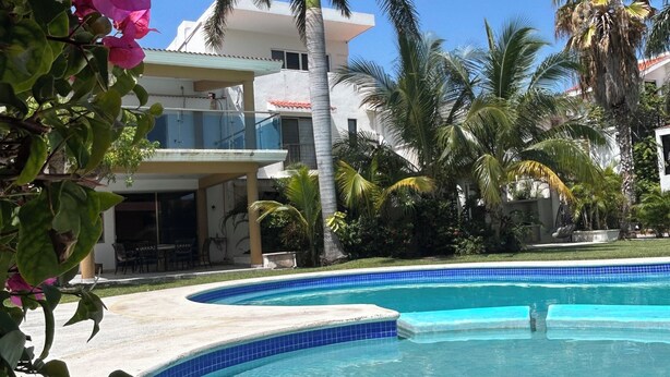 A swimming pool with clear blue water, surrounded by palm trees and a building with balconies. - Cancun House in Hotel Zone (Cancún)
