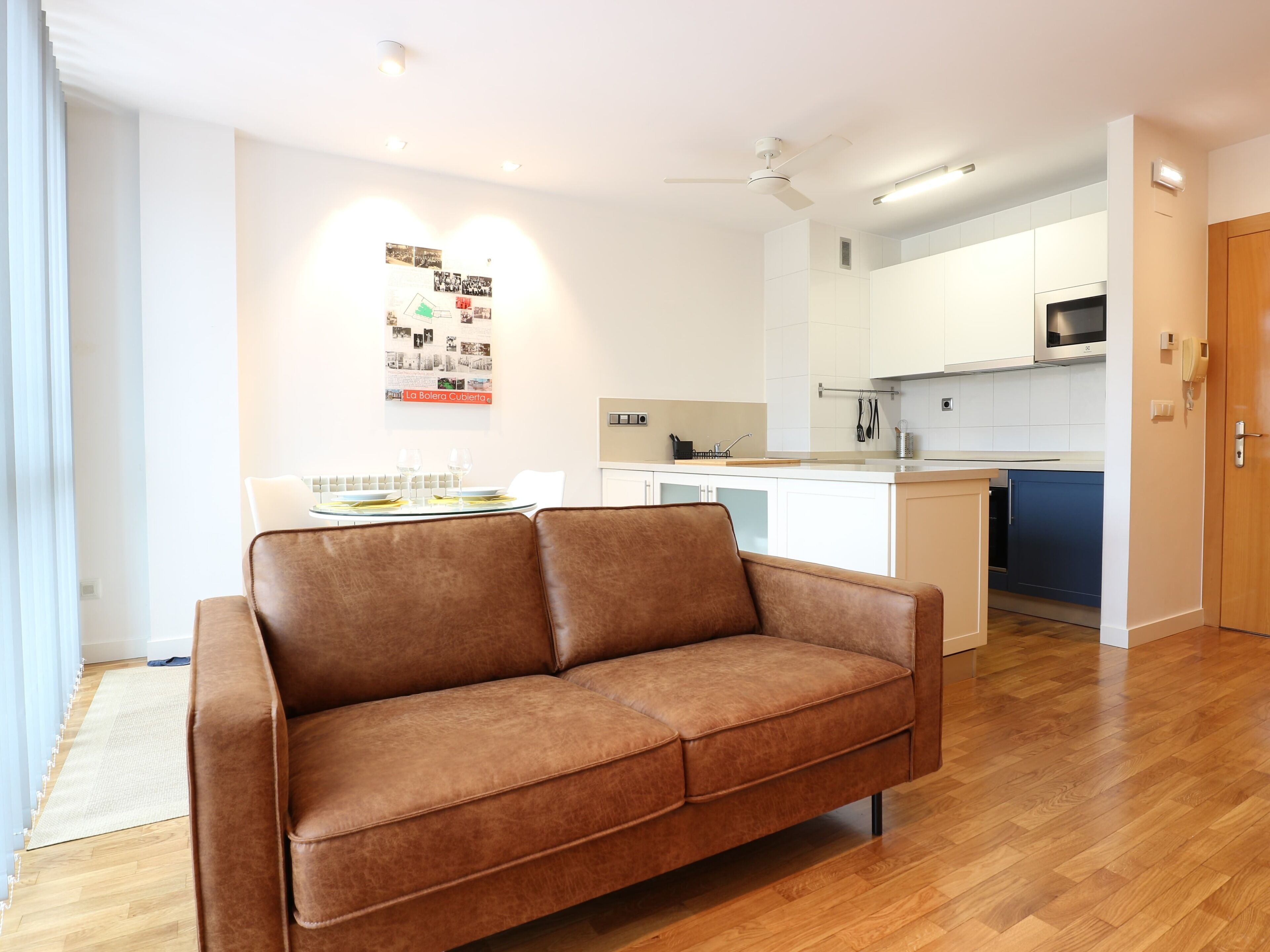 Una sala de estar moderna con un sofá de cuero marrón, una mesa de comedor con dos vasos, y una cocina al fondo.