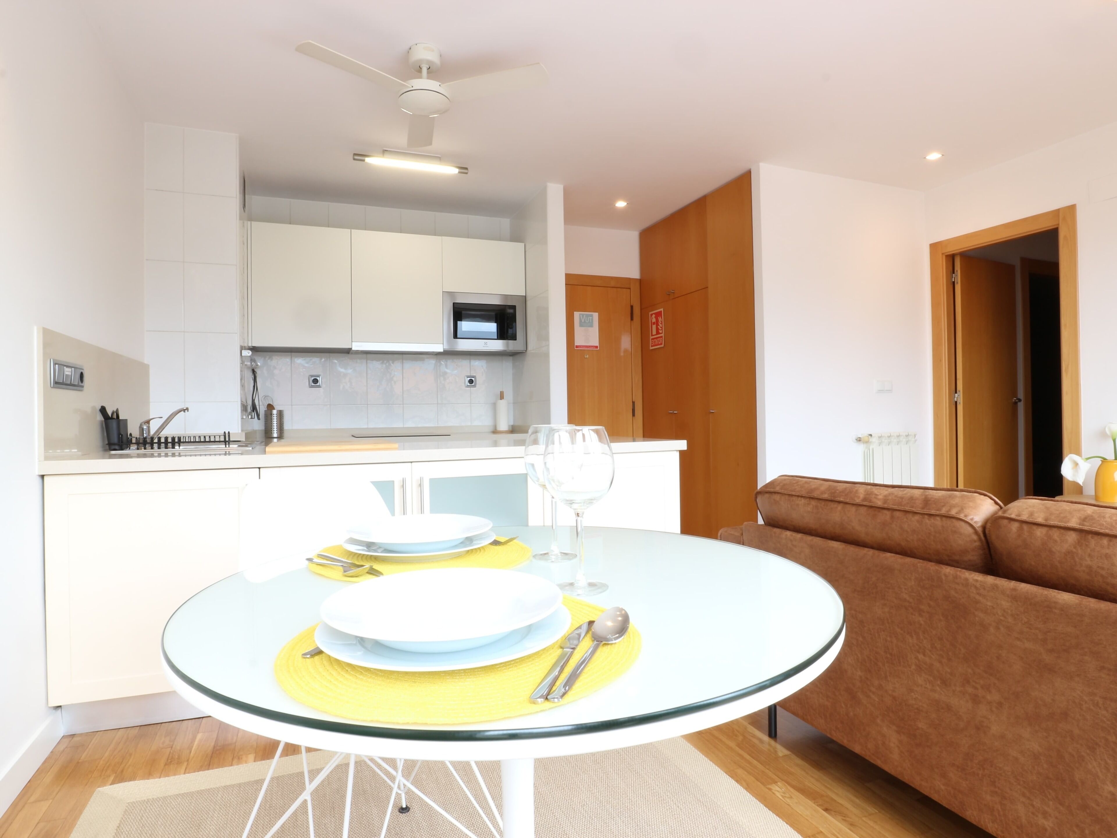 Una cocina moderna con armarios blancos, microondas y un comedor con una mesa redonda de vidrio para dos.