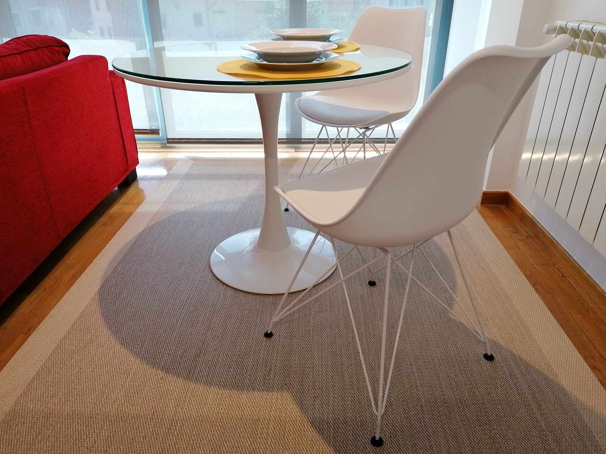 A modern living room with a glass-top coffee table, white chairs, a red sofa, and a patterned rug.